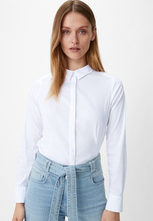 Camicia bianca con colletto, maniche lunghe e polsini; abbinata a jeans a vita alta azzurri con una cintura legata.