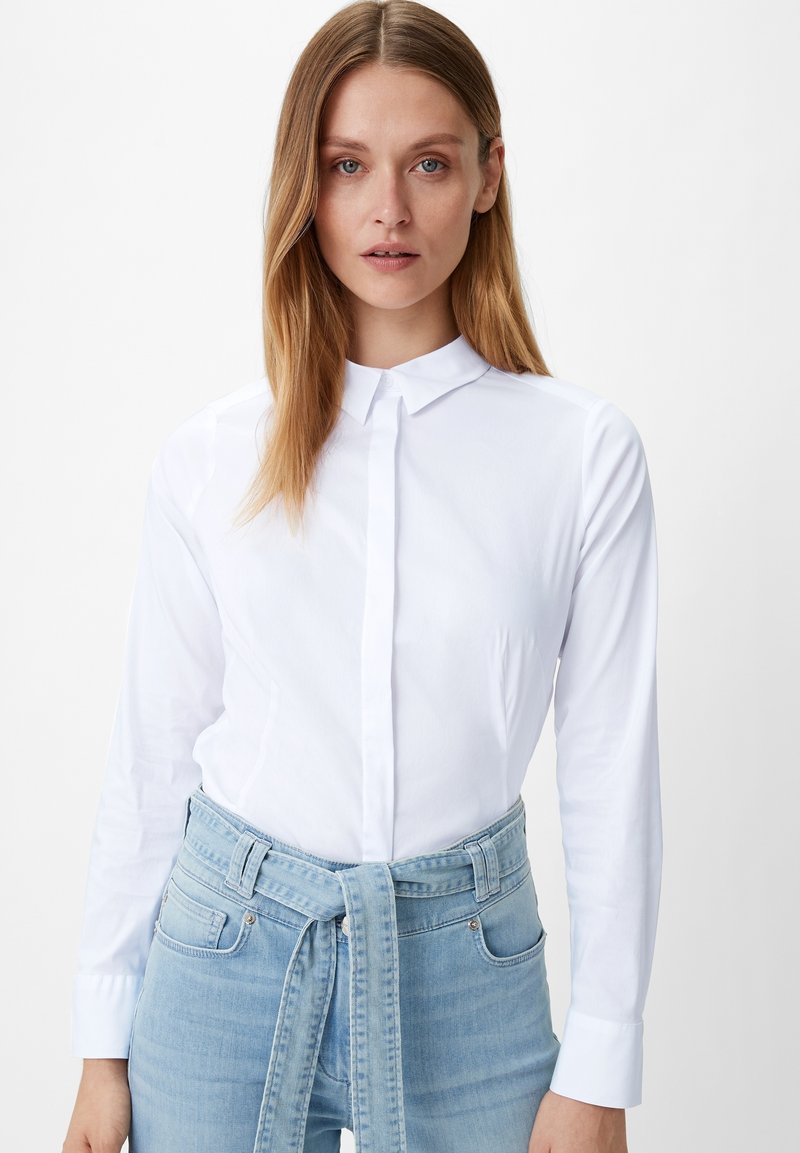 Camicia bianca con colletto, maniche lunghe e polsini; abbinata a jeans a vita alta azzurri con una cintura legata.
