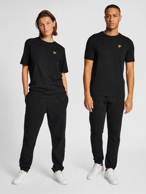 Twee modellen dragen zwarte bijpassende casual outfits met kleine gele logo's en witte sneakers, staand tegen een effen achtergrond.