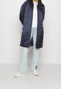 Marine satin lang jakke med lynlås foran, parret med en hvid sweatshirt og lyseblå flared jeans. Hvide sneakers fuldender outfittet.