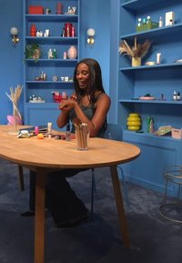 Runt träbord med blå ben, täckt med olika kosmetika och verktyg. Blå bakgrund med hyllor som visar färgglada produkter.