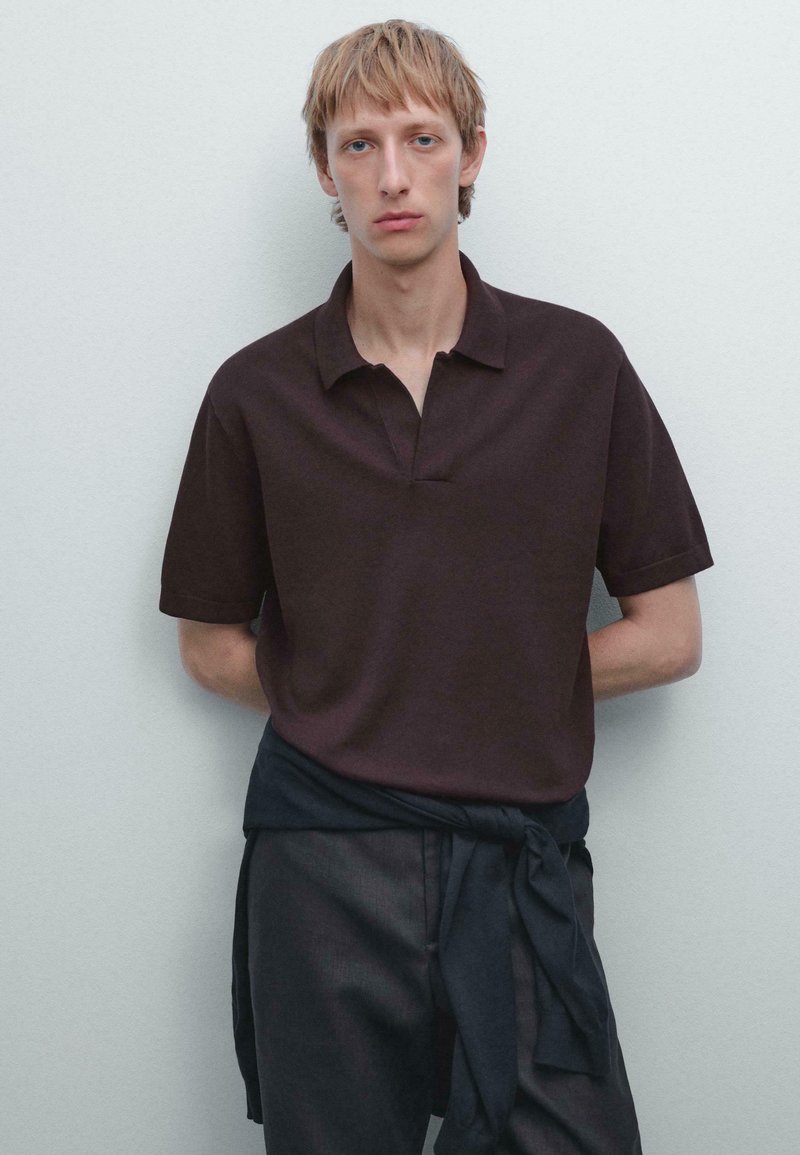 Brun polo-shirt med kort krave og korte ærmer, parret med mørke bukser og et sort bælte om livet. Neutral baggrund.