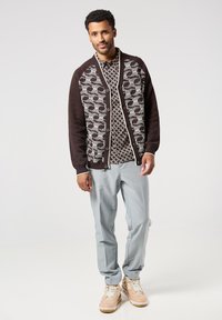 Tmavě hnědý cardigan s bílým geometrickým vzorem, nošený přes vzorovanou košili. Světle šedé kalhoty a béžové tenisky doplňují outfit.