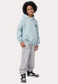 Felpa in pile azzurro chiaro con tasca frontale e testo "STAY AWESOME", abbinata a pantaloni cargo grigio chiaro con più tasche e sneakers nere.