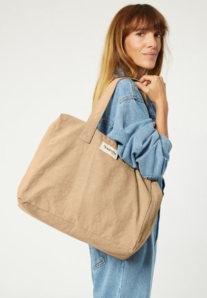 Beige Canvas-Einkaufsbeutel mit stabilen, flachen Griffen und einer weichen Textur; verfügt über ein sichtbares Markenschild auf einer Seite.