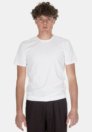 Giovane uomo con capelli ricci che indossa una maglietta bianca semplice a maniche corte e pantaloni neri, in piedi davanti a uno sfondo bianco.