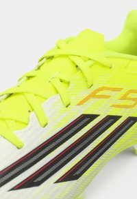 Λαμπερό κίτρινο ποδοσφαιρικό παπούτσι με μαύρες και λευκές λεπτομέρειες, διαθέτοντας υφή στην επάνω επιφάνεια και έντονο σύστημα κορδονιών. Διακριτικός σχεδιασμός με λωρίδες.