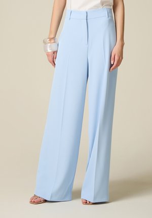 Vrouw draagt lichtblauwe broek met wijde pijpen, wit topje, doorzichtige armbanden en open sandalen, staand tegen een neutrale achtergrond.