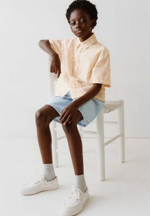 Jeune personne portant une chemise claire à motifs, un short en jean, des chaussettes grises et des baskets blanches, assise avec un bras reposant sur une chaise blanche.