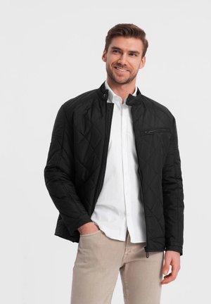 Black quilted jacket with a high collar and zippered front. Features a zippered chest pocket and smooth fabric texture. Paired with a white shirt.