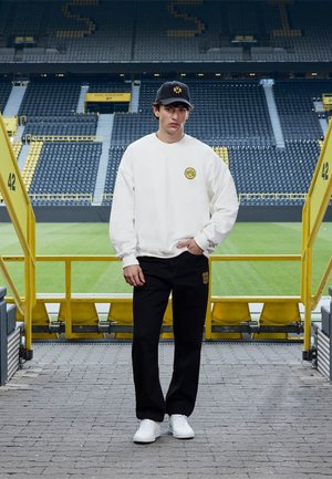 Jeune homme portant un sweat-shirt blanc et un pantalon noir avec les logos du Borussia Dortmund se tient dans un stade de football vide.