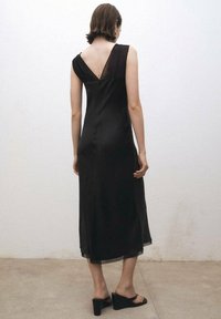 Schwarzes, ärmelloses Kleid mit V-Ausschnitt, knielanger Saum und glatter Textur, das ein tiefes Rückendesign aufweist. Getragen mit schwarzen Keilsandalen.