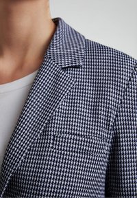 Blazer à motif pied-de-poule bleu marine et blanc avec un col cranté, une unique poche poitrine et un design d'épaule structuré. Texture de tissu douce.