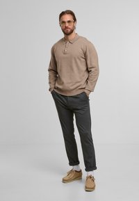 Man in een beige lange mouw polo, donkergrijze opgerolde broek, witte sokken en beige suède schoenen, staande met zijn handen in zijn zakken tegen een grijze achtergrond.