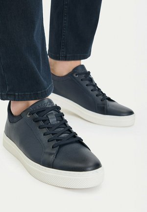 Person wearing black leather sneakers with white soles and black laces, paired with dark ankle-length jeans.