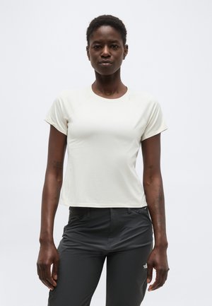 Vrouw met kort haar, gekleed in een nauwsluitend wit shirt met korte mouwen en een donkere broek, staand tegen een effen lichte achtergrond.