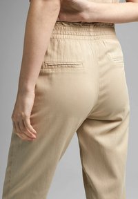 Personne portant un pantalon beige taille haute avec une ceinture élastique, montrant les poches arrière, sur un fond gris uni.