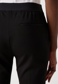 Černé kalhoty vyrobené z jemné, hladké tkaniny. Pas je elastický, má diskrétní, elegantní zadní kapsu a minimální detaily.