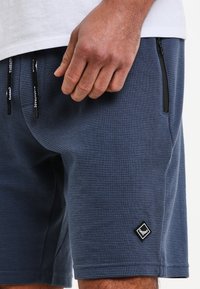 Námořnické modré šortky vyrobené z texturované tkaniny, které mají černé kapsy na zip a malou černou nášivku ve tvaru diamantu na boku.