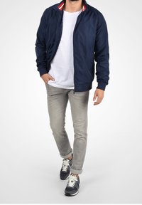 Navy bomber jacket with white and red striped collar, worn over a white t-shirt, paired with light gray jeans and dark sneakers.