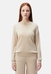 Beige stickad tröja med långa ärmar och rundad hals. Mjuk textur och ribbad fåll. I kombination med matchande beiga byxor för att skapa en sammanhängande look.