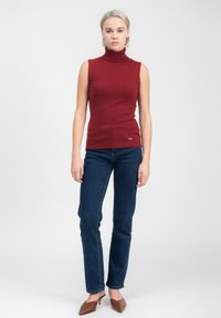 Maglione senza maniche rosso con collo a dolcevita e texture a coste, abbinato a jeans blu scuro a gamba dritta e mule con tacco marroni.