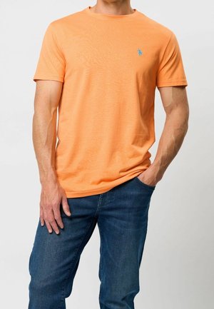 Man draagt een effen oranje T-shirt met korte mouwen en blauwe jeans, met één hand in de zak tegen een effen lichte achtergrond.