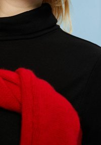 Schwarzer Rollkragenpullover mit gerippter Textur, ergänzt durch einen gefalteten roten Strickschal, der über die Schulter drapiert ist.