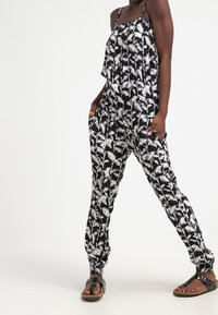 Svart och vit jumpsuit med palmbladsmönster, spagettiband, elastisk midja och fickor; bärs med avslappnade sandaler.