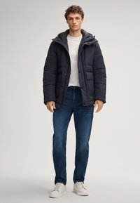 Mann in einer dunkelblauen Steppjacke über einem weißen Hemd, blauen Jeans und weißen Sneakers, vor einem schlichten Hintergrund stehend.