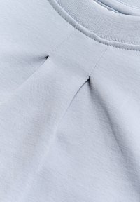 Heled halli puuvillane kangas, millel on tekstuuriline pind, kaeluse lähedal pleedi detail ja soonikuga krae.