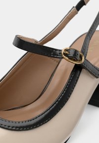 Chaussure beige avec une bordure noire et une fine sangle noire maintenue par une petite boucle ronde dorée.