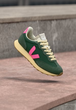 Zielony sneaker z różowym logo "V", kremowymi sznurowadłami i beżową podeszwą, unoszący się nad różową kamienną powierzchnią i szarym kamiennym tłem.