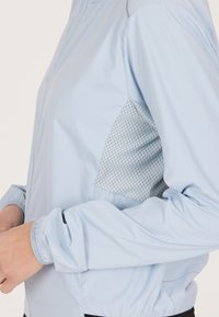 Hellblaues Jäckchen aus glattem Stoff mit elastischen Bündchen und einem strukturierten Mesh-Einsatz an der Seite für Atmungsaktivität. Leicht und tailliert.