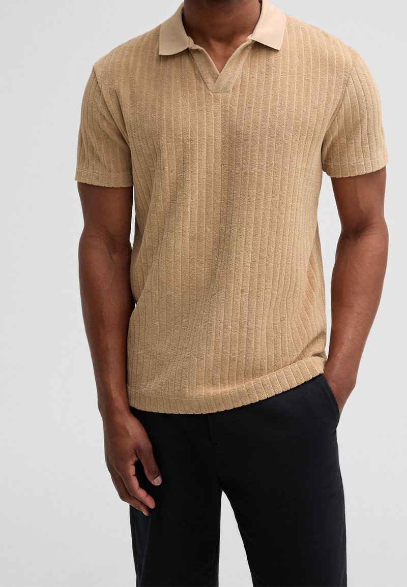 Homme portant un polo beige à manches courtes en maille côtelée avec un pantalon noir, debout avec une main dans la poche sur un fond uni.