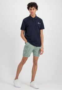 Polo shirt blu navy con colletto e tre bottoni; abbinato a pantaloni cargo verdi con tasche laterali; scarpe da ginnastica bianche completano l'outfit.