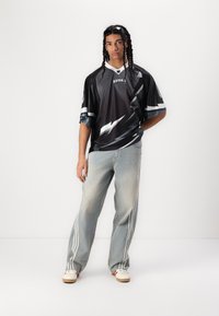 Maillot de sport noir avec des rayures blanches en forme d'éclair, coupe ample, associé à un jean en denim clair avec trois rayures sur le côté et des chaussures blanches.