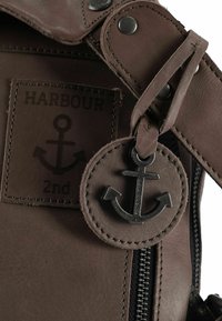 Bruine leren tas met een getextureerde afwerking, voorzien van een ritssluiting, metalen beslag en een aan het hengsel bevestigde tag in de vorm van een anker.