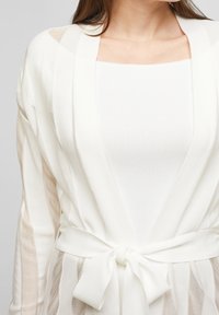 Biały kardigan wykonany z lekkiego, półprzezroczystego materiału. Posiada wiązanie w talii, otwarty przód oraz długie rękawy z pionowymi paskami.