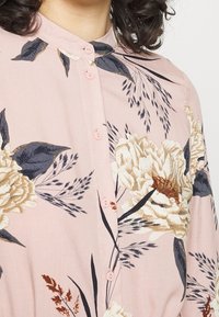Žena v bledě růžové košili s knoflíky, pokryté velkými krémovými a hnědými květinovými vzory a tmavými listy, viditelné kudrnaté tmavé vlasy.