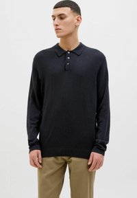 Svart langermet polo skjorte med krage, tre-knappers placket og ribbede mansjetter; brukt med khaki-bukser; myk tekstur.