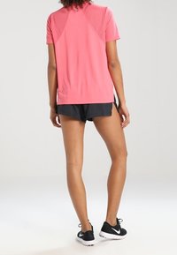 Kobieta w różowej, krótkiej koszulce sportowej, czarnych szortach do biegania i czarnych sneakersach Nike, stojąca tyłem, zwrócona do przodu.