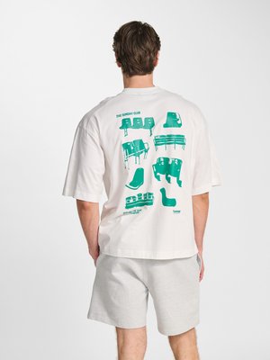 Man draagt een wit oversized T-shirt met groene stadionzitplaatsgrafieken en grijze korte broek, staand met de rug naar de camera.