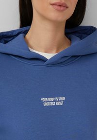 Blauwe hooded sweatshirt van zachte stof met een witte tekstprint die zegt: "JOUW LICHAAM IS JE GROTE TROEF."