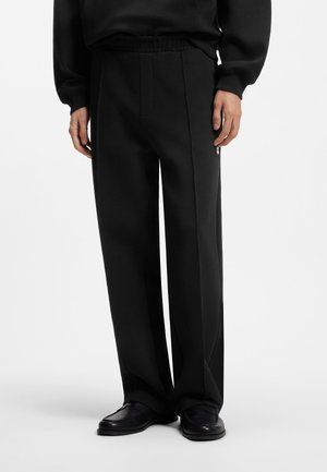 Personne portant un pantalon noir ample avec un pli à l'avant, une taille élastique et des chaussures en cuir noires sur un fond uni.