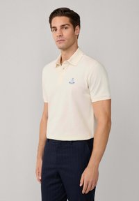 Cremfarbendes Poloshirt mit einer Knopfleiste mit zwei Knöpfen, kurzen Ärmeln und einem gestickten blauen Logo auf der linken Brust. Kombiniert mit nautisch gestreiften Stoffhosen in Marineblau.