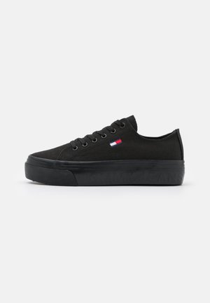 Czarny niskoprofilowy sneaker ze sznurowadłami, grubą gumową podeszwą z wytłoczonym nazwą marki i małym logo flagi w kolorach czerwonym, białym i niebieskim po boku.