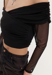 Czarna bluzka na ramiączkach z marszczonym przodem, przezroczystymi długimi rękawami oraz gładką strukturą. W zestawie z ciemnymi dżinsami z wysokim stanem.
