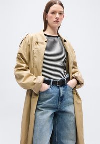 Γυναίκα με ίσια καστανά μαλλιά φοράει ένα μπεζ παλτό-καπαρντίνα, μαύρη και λευκή ριγέ μπλούζα, τζιν παντελόνι ψηλόμεσο μπλε και μαύρη ζώνη με τα χέρια στις τσέπες.