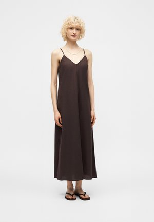 Szczupła osoba z kręconymi blond włosami, ubrana w bezrękawnikową ciemnobrązową sukienkę maxi i czarne japonki, stoi na jednolitym tle.